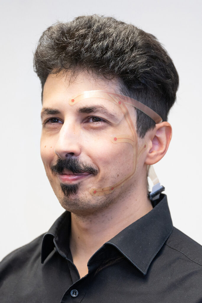 Porträt einer Person mit kurzen dunklen Haaren. Auf Stirn, Schläfe und Wange ist schematisch ein transparentes EEG-Sensorpatch mit markierten Kontaktpunkten dargestellt, das die Position der Elektroden zur Erfassung von Hirnsignalen zeigt.