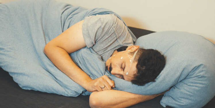 Eine Person liegt schlafend auf der Seite in einem Bett und ist zugedeckt. Am Kopf sind mehrere kleine Sensoren angebracht, die über feine Kabel mit einem kompakten Messgerät am Hals verbunden sind. Die Sensoren erfassen Hirnaktivität und weitere Biosignale während des Schlafs. Die Person befindet sich in einer entspannten Schlafposition in einer ruhigen, häuslichen Umgebung. Das Bild veranschaulicht eine mobile EEG-Messung unter Alltagsbedingungen.