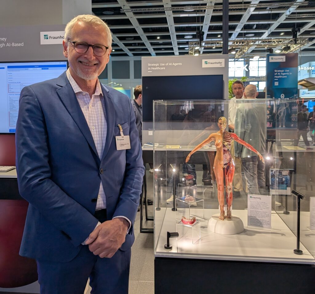 Ein Mann im Anzug steht auf einer Messe neben einem gläsernen Vitrinenkasten, in dem ein medizinisches Modell eines menschlichen Körpers ausgestellt ist.