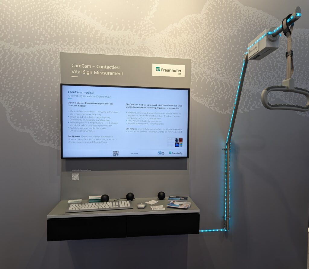 Ein Monitor ist mit einem Gestell verbunden, welches mit einem Kamerasystem ausgestattet ist. Ein Griff hängt nach unten (symbolisiert Krankenbett)