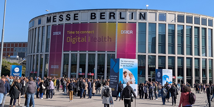 Ein großes Gebäude mit Glasscheiben und dem Schrifzug "Messe Berlin". Menschen strömen zum Eingang, der Himmel ist blau.