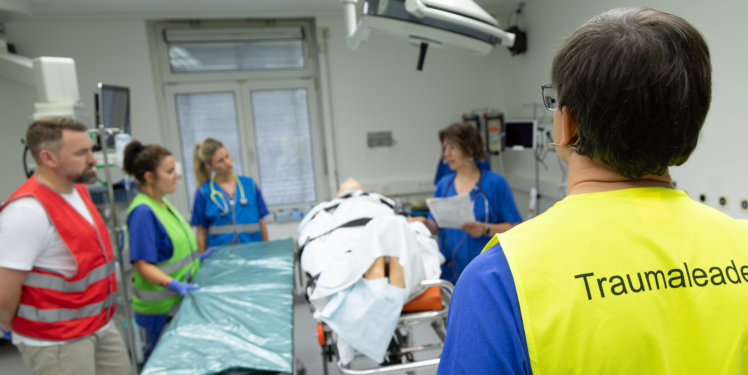 Ein medizinisches Team bespricht sich in einem Schockraum rund um eine zugedeckte Person auf einer Patientenliege. Im Vordergrund steht eine Person mit gelber Warnweste und der Aufschrift „Traumaleader“.